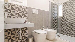 A modern bathroom with a geometric tile backsplash, a white sink, two toilets, and a glass-enclosed shower. - hilde's house 2 (Scicli)