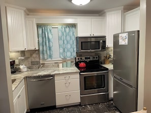 Una cocina con armarios blancos, un microondas de acero inoxidable, una estufa, un refrigerador y un ventanal con cortinas estampadas.