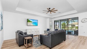 A spacious living room with a large gray sofa, a wooden coffee table, and a TV mounted on the wall. There is a ceiling fan, a window with a view of the outdoors, and a clock on the wall. - Great Location! 2,444 sq ft canal front villa with heated pool/spa. (Cape Coral)