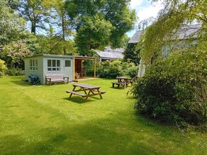 A small wooden cabin with a porch, a picnic table, and a shed in a lush green garden. - A charming and spacious cottage in rural St Ives (Cornwall)