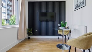 A modern living room with a flat-screen TV mounted on a dark wall, a small round table with a chair, a potted plant, and a window with curtains. - Cosy Studio Apartment in Canary Wharf (London)