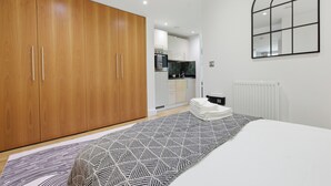A bedroom with a bed, a geometric-patterned rug, wooden wardrobe, and a kitchen in the background. - Cosy Studio Apartment in Canary Wharf (London)
