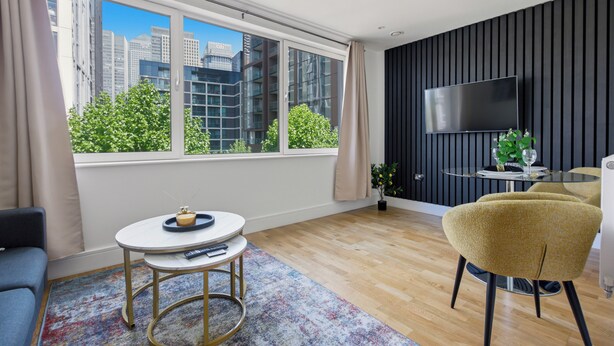 A modern living room with a large window, a flat-screen TV, a round table, a sofa, and a yellow armchair. - Cosy Studio Apartment in Canary Wharf (London)