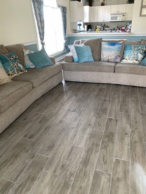 A living room with a sectional sofa, a kitchen in the background, and wooden flooring.