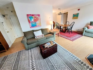 Un salon comprenant un canapé, une table basse, un tapis à motifs, un coin repas avec une table et des chaises, et une peinture abstraite aux couleurs vives accrochée au mur.