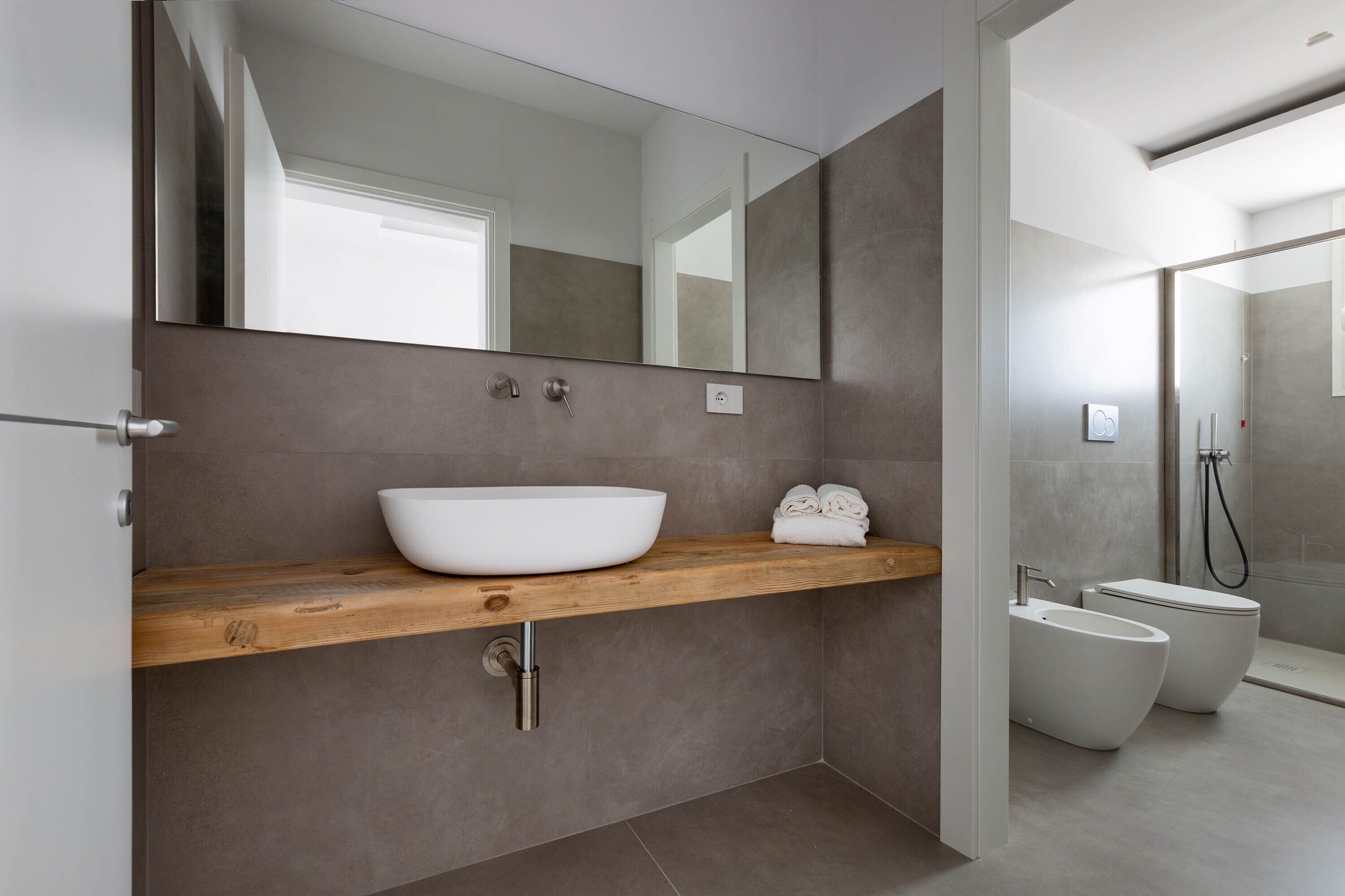 Une salle de bain moderne avec un grand miroir, un meuble-lavabo en bois, un lavabo blanc, deux toilettes et un espace douche.