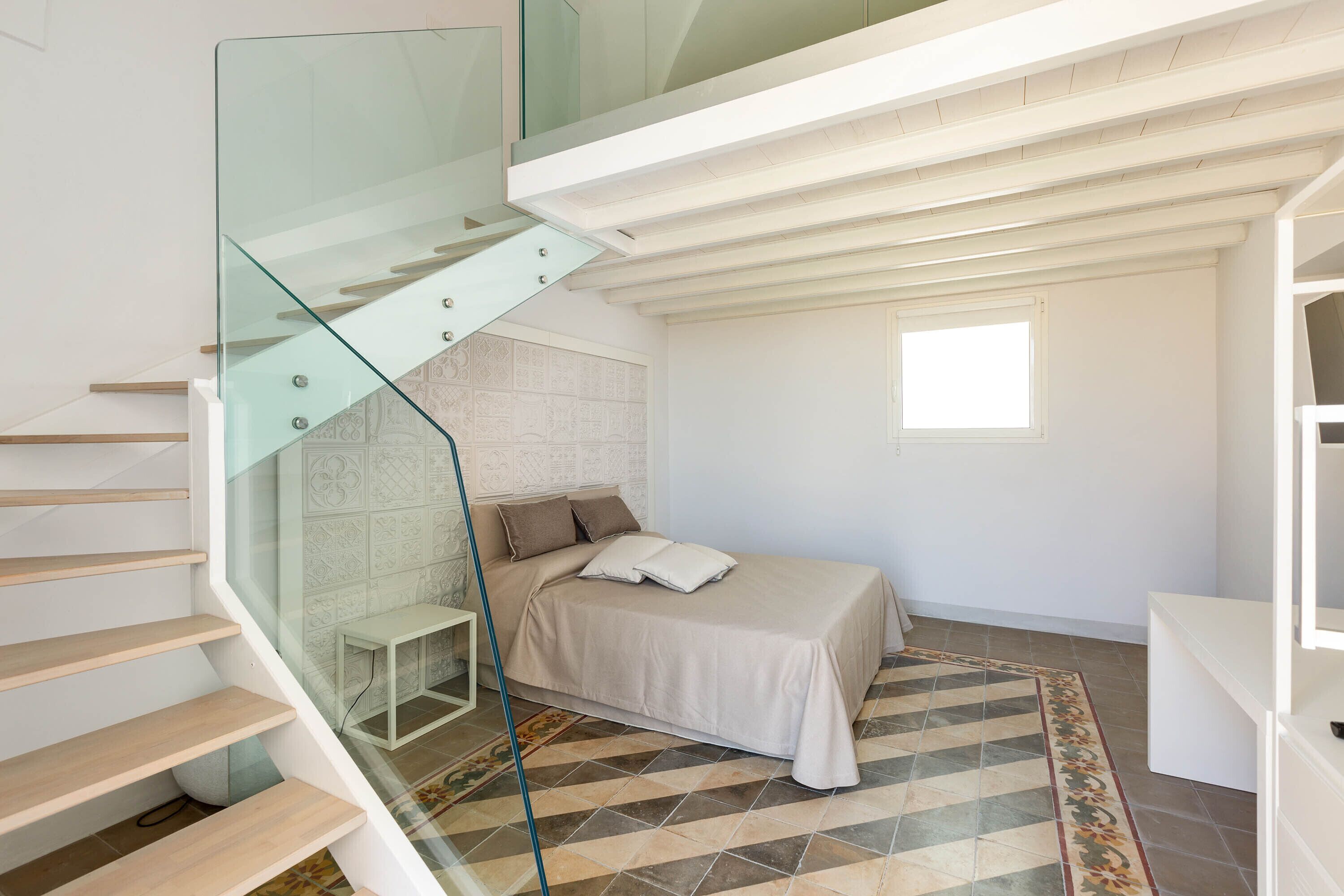 A bedroom with a bed, a glass staircase, a small table, and a patterned rug.