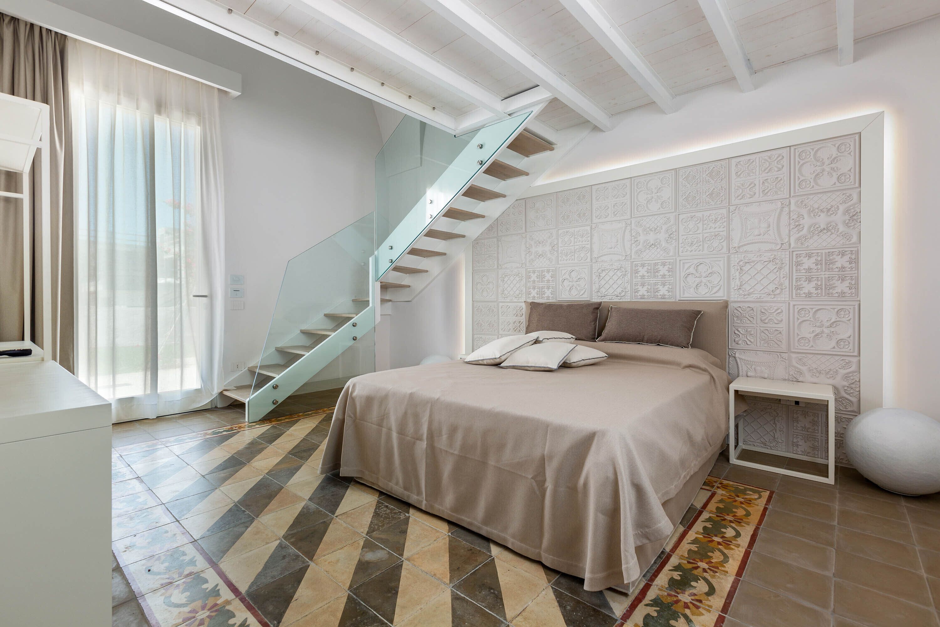 A bedroom with a bed, a glass staircase, a patterned wall, and a tiled floor.