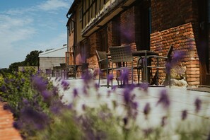 Un bâtiment de style rustique, doté d’une façade en briques, d’un espace extérieur aménagé pour s’asseoir et d’un jardin où poussent des fleurs violettes.