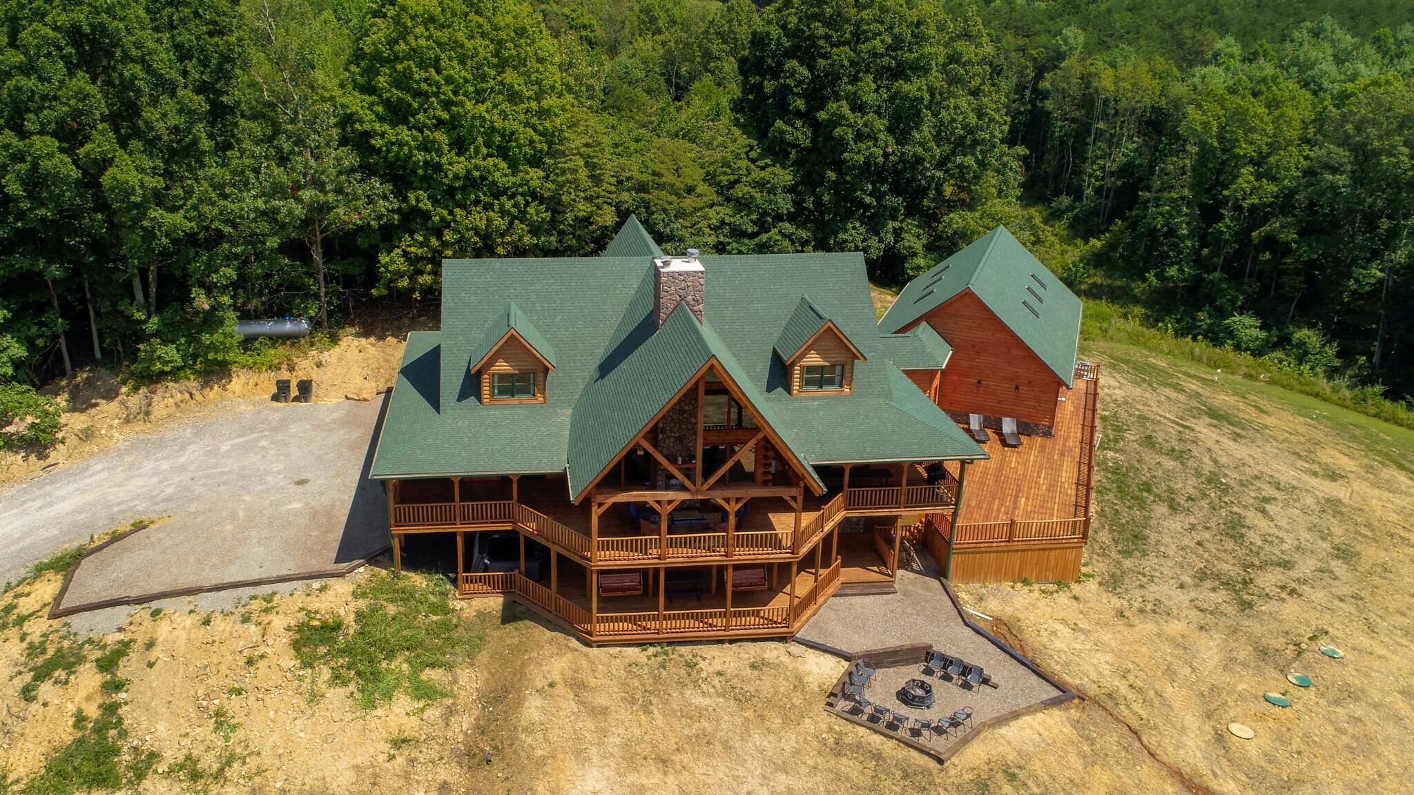 A large log cabin with multiple decks and a stone chimney, surrounded by dense forest.