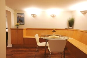 Un pequeño comedor con una mesa redonda de vidrio, dos sillas blancas, un banco de madera, un armario y una lámpara fijada a la pared.