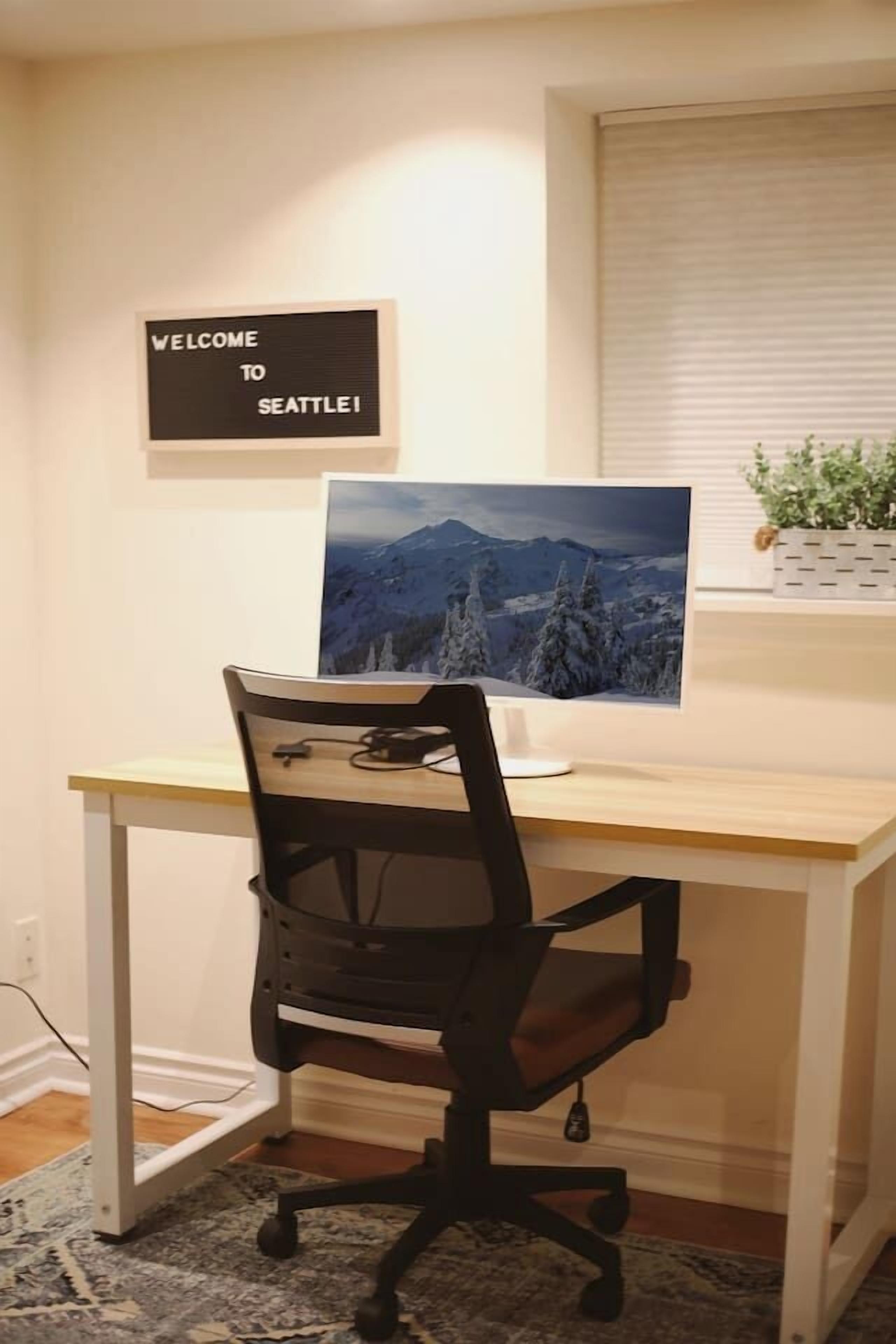 Un espace de travail comprenant un bureau, un ordinateur et une chaise, une pancarte « Bienvenue à Seattle » et une photographie représentant un paysage de montagne.