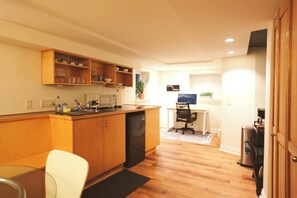 Cocina compacta con armarios de madera, microondas y pileta. Hay una zona de comedor con una silla blanca y una mesita. Al fondo, un escritorio con una silla.