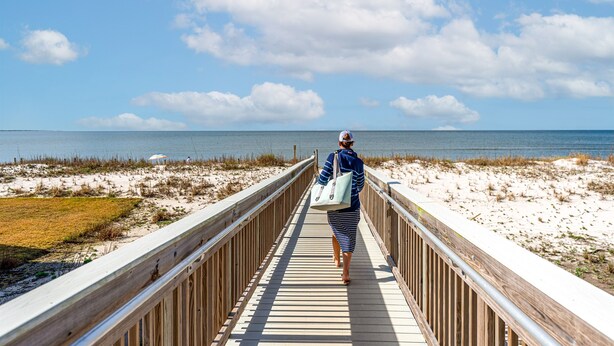 En træpromenade der fører til en strand med en klar himmel og hav i baggrunden.