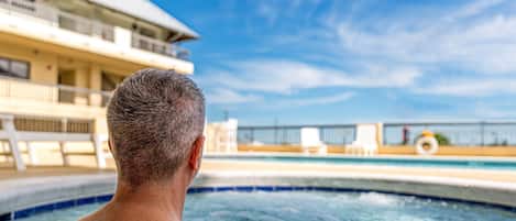 A man in a hot tub with a view of a pool and a building in the background.