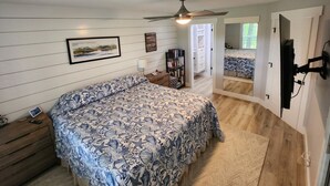 A bedroom with a bed, a ceiling fan, a TV, and a door leading to another room. - Modern Beach Elegance: The Maples Retreat (Sunset Beach)