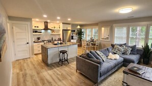 A modern kitchen with a central island, a dining area with a table and chairs, a grey sofa, and a pet dog. - Modern Beach Elegance: The Maples Retreat (Sunset Beach)