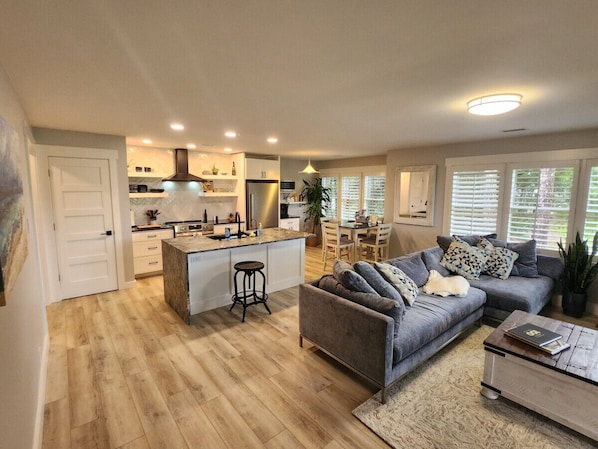Una cocina moderna con una isla central, un área de comedor con mesa y sillas, un sofá gris y un perro mascota.