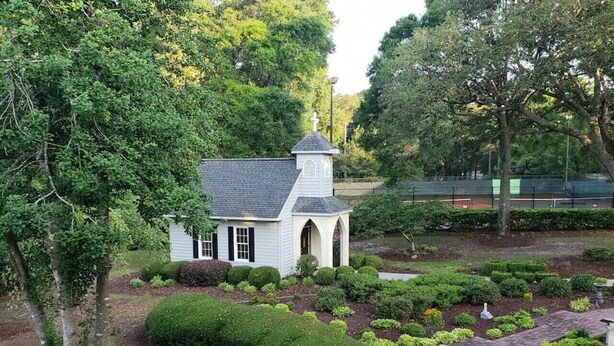 A small white chapel with a pointed roof and a cross on top, surrounded by well-manicured gardens and hedges.