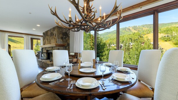 Ein Esszimmer mit einem großen runden Tisch, gedeckt für sechs Personen, umgeben von komfortablen Stühlen, einem Kamin aus Stein und Blick auf die Berge.
