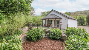 A small white building with a porch, surrounded by lush greenery and a wooden deck.