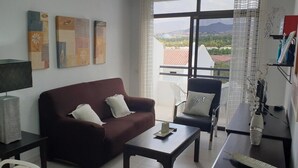 Ett vardagsrum med en soffa, ett soffbord och en fåtölj. På väggen hänger tre tavlor, och det finns en lampa. Genom ett fönster kan man se ut.