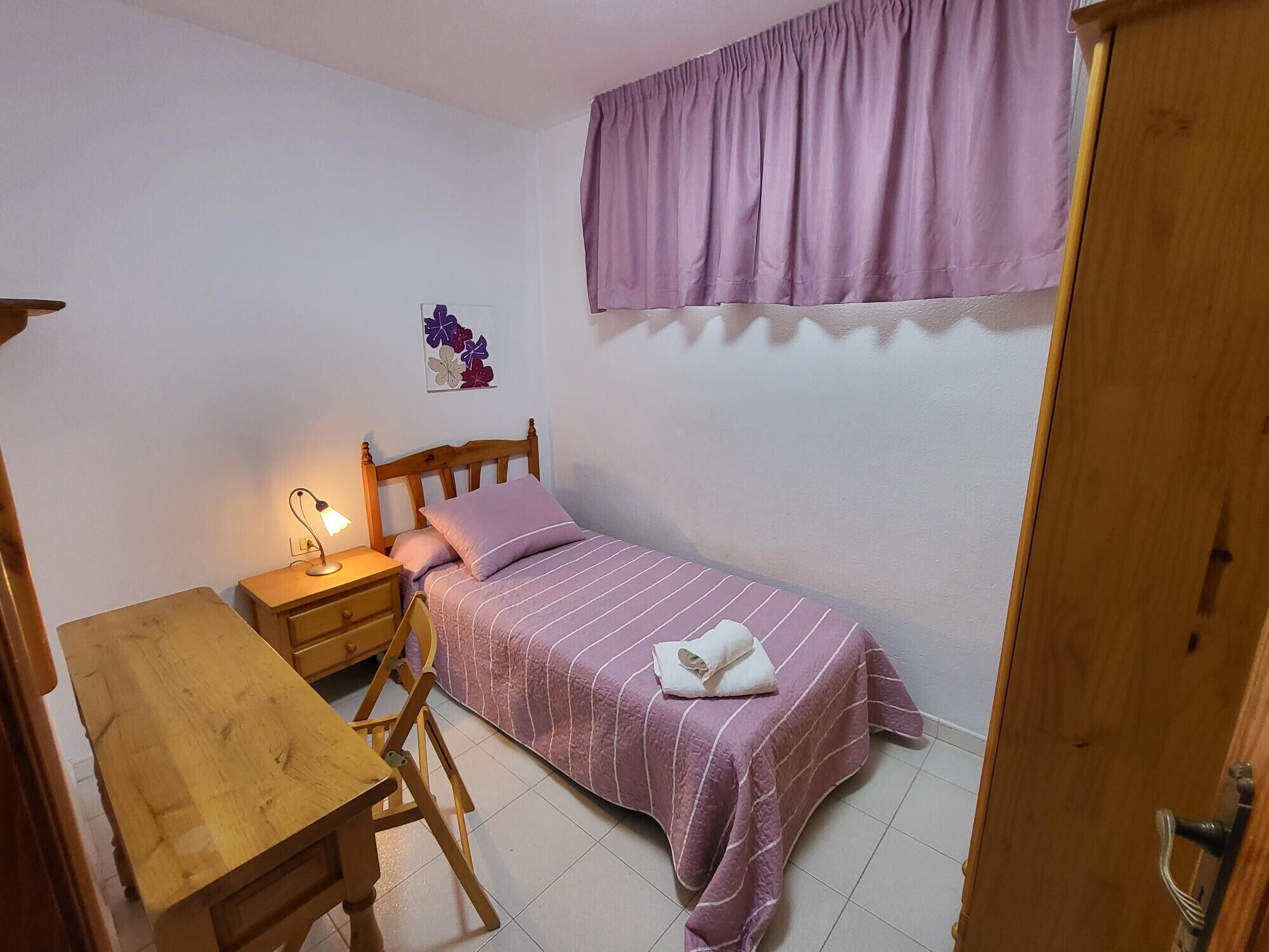 A small bedroom with a single bed, a wooden nightstand, a table, and a wall-mounted lamp.