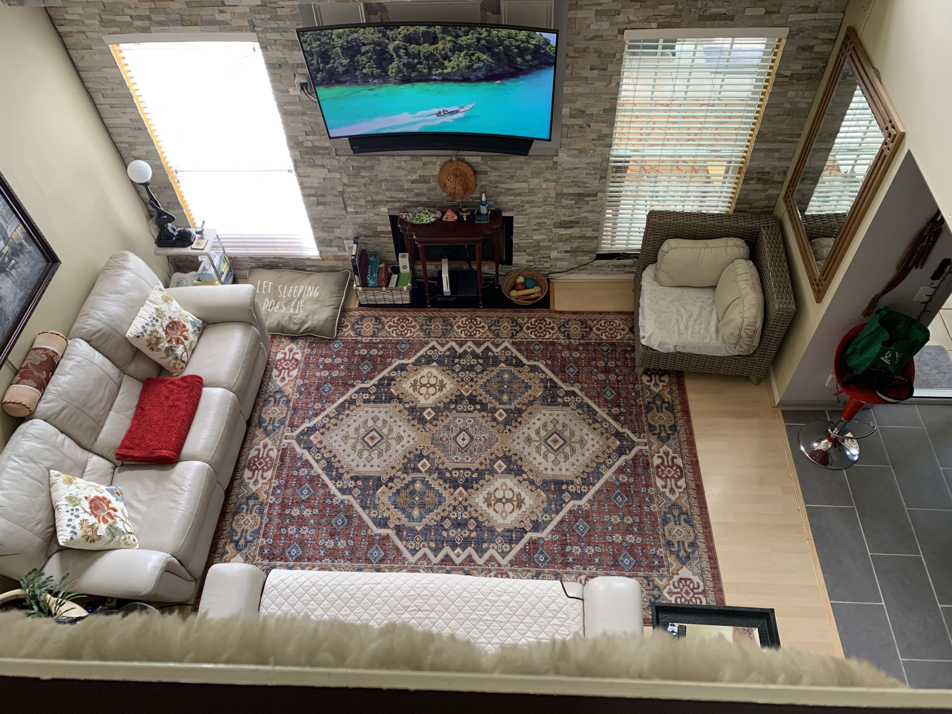 Un salon doté d’un grand téléviseur à écran plat fixé sur un mur en pierre, d’un tapis à motifs, d’un canapé agrémenté de coussins décoratifs et d’un fauteuil en osier.