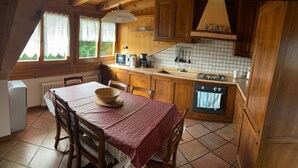 Une cuisine chaleureuse avec des meubles en bois, un pare-feu en carrelage, une table à manger recouverte d’une nappe rouge et une fenêtre ornée de rideaux en dentelle.