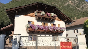 Un chalet traditionnel doté d’un balcon en bois orné de fleurs, d’une entrée fermée par un portail et d’une enseigne indiquant « Appartements Giustina ».