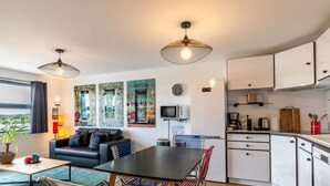 A modern living room with a black sofa, a dining table, and a kitchen area with white cabinets and appliances. - L'Annexe du Cosmopolitain | Luxury Building | 4 Guests (Honfleur)