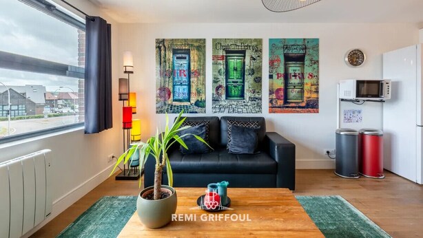 A modern living room with a black sofa, a wooden coffee table, and a potted plant. There are colorful abstract paintings on the wall and a window with a view of buildings. - L'Annexe du Cosmopolitain | Luxury Building | 4 Guests (Honfleur)