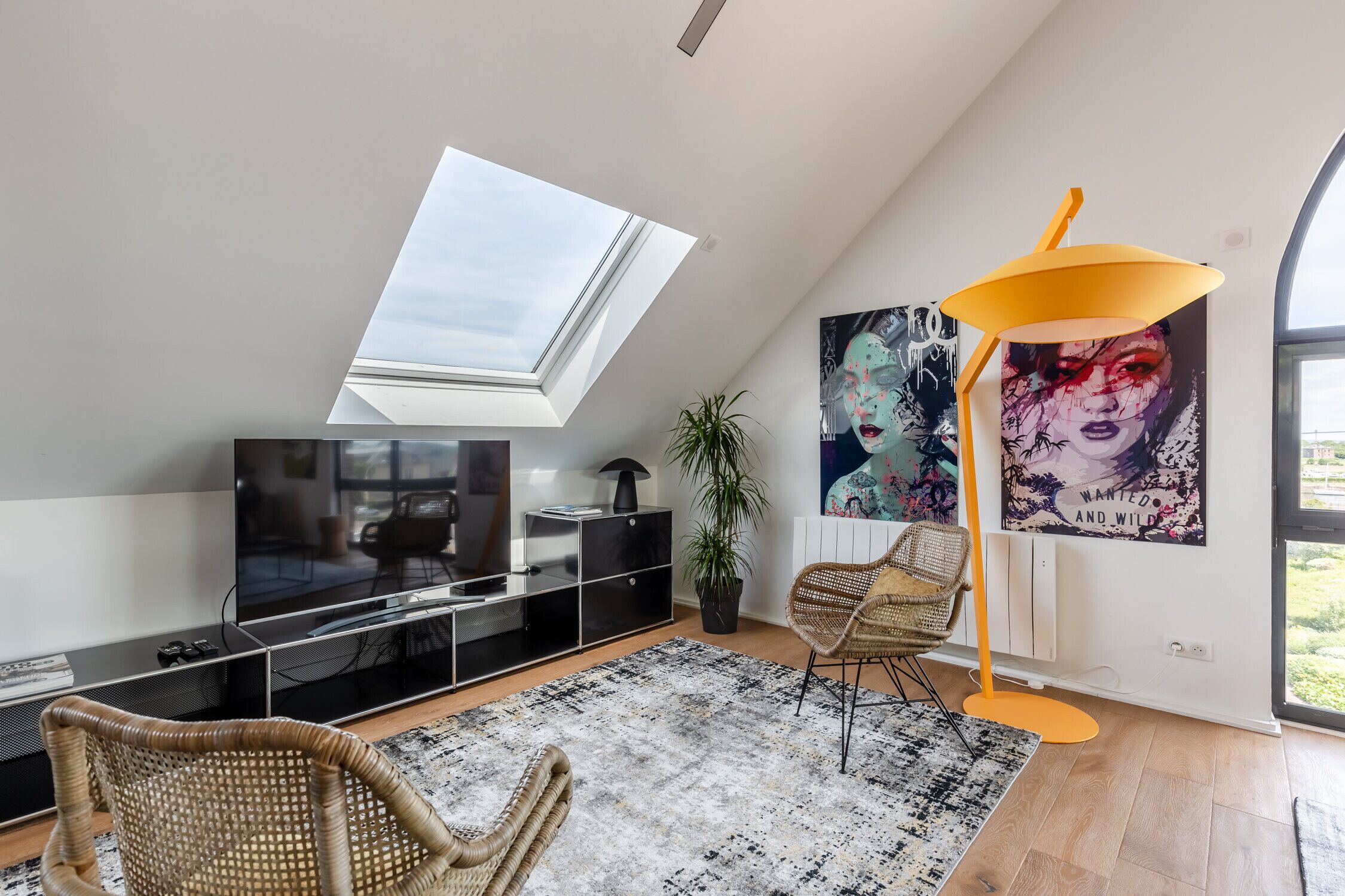 Una sala de estar moderna con tragaluz, un televisor plano grande, un mueble de entretenimiento negro, una lámpara de pie amarilla, una alfombra blanco y negro, y un cartel en la pared.