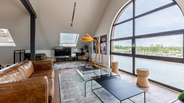 Un salon spacieux avec un canapé en cuir, une table basse en verre et une grande fenêtre donnant sur une étendue d’eau.