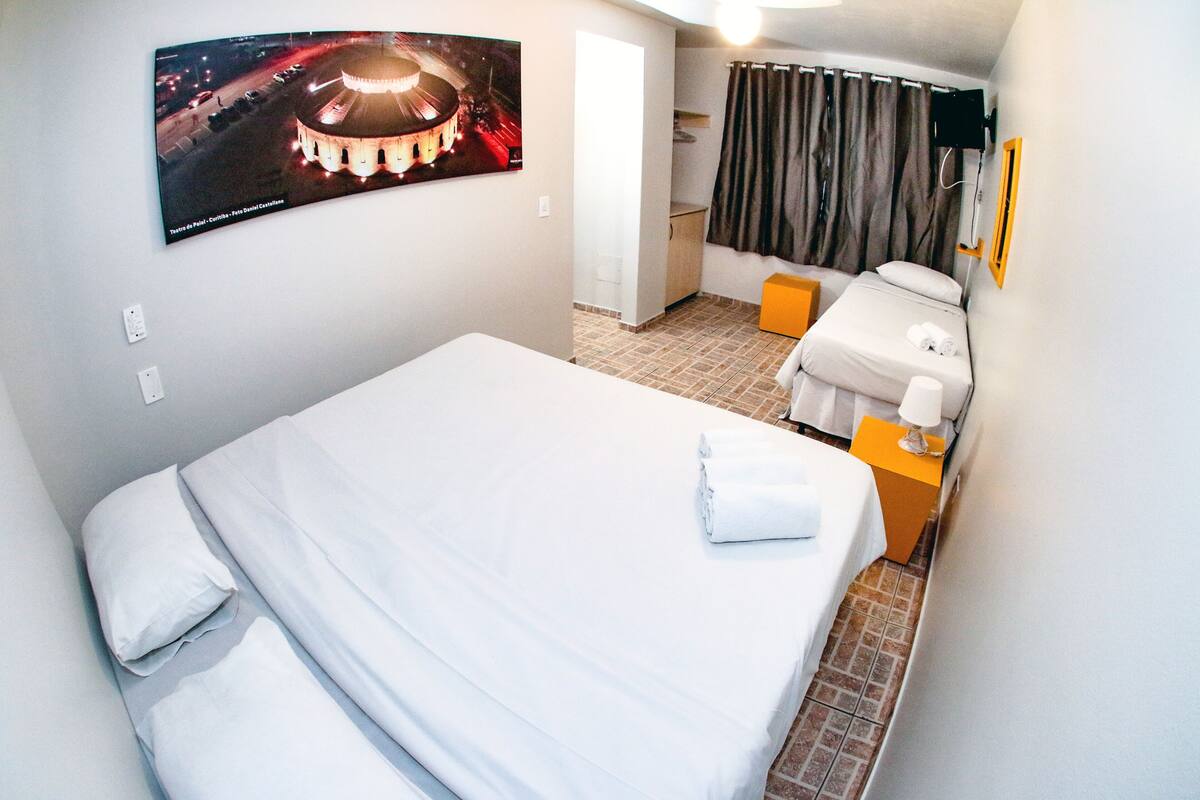 A hotel room with a bed, a small table with a lamp, a bathroom with a shower curtain, and a poster on the wall.