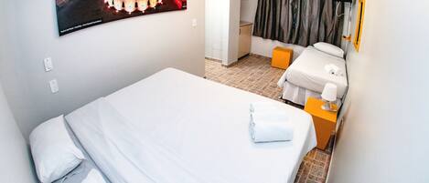 A hotel room with a bed, a small table with a lamp, a bathroom with a shower curtain, and a poster on the wall.