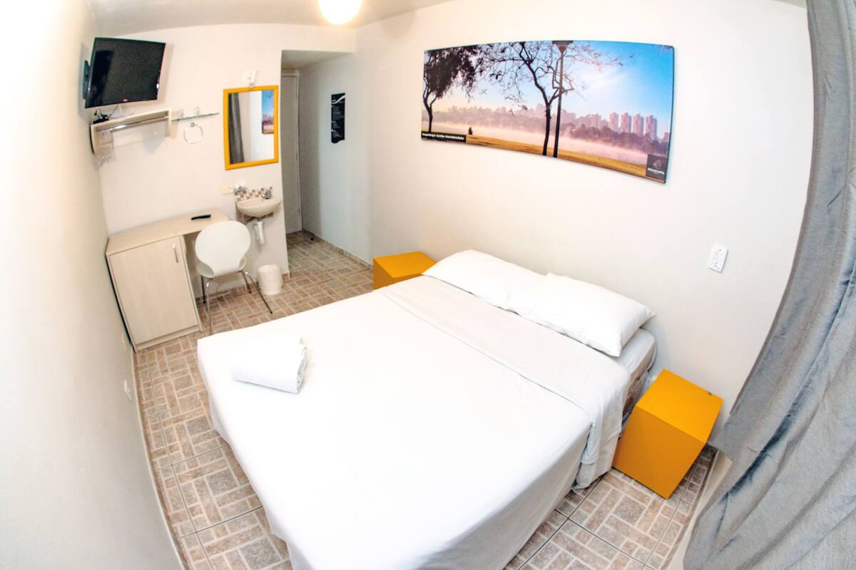 A hotel room with a bed, a bathroom, a TV, and a cityscape painting.