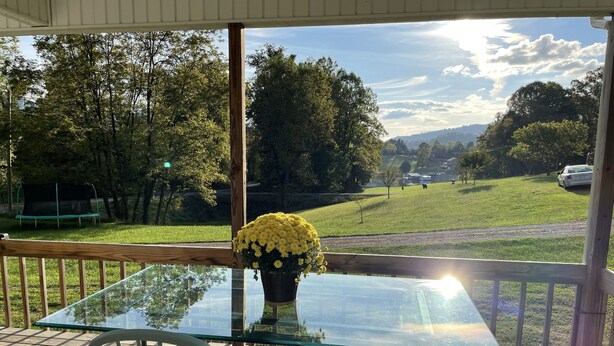 Ein Glastisch mit zwei Stühlen auf einer Holzterrasse, mit Blick auf eine Rasenfläche mit Bäumen und eine entfernte Bergsicht.