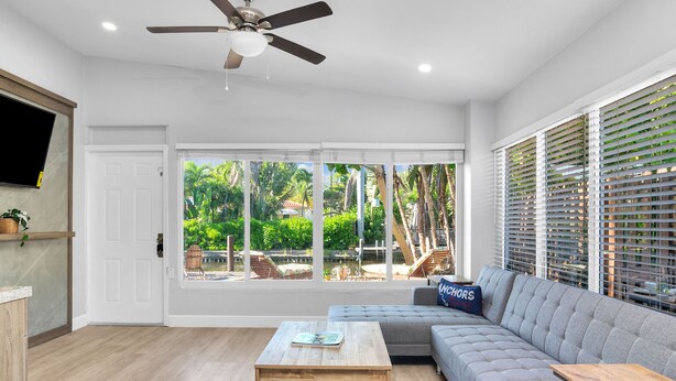 A spacious living room with a large window, a ceiling fan, a grey sofa, and a wooden coffee table. - Riverside villa next to Down Town Ft Lauderdale (Fort Lauderdale)