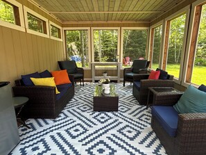 A covered patio area with wicker furniture, a patterned floor, and large windows offering a view of the outdoors. - Modern Vacation Home With A/C & Screened In Room. Close To National Park Beach (Brackley Beach)