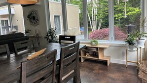 Un coin repas avec une table et des chaises en bois, de grandes fenêtres donnant sur un paysage verdoyant et un luminaire suspendu.