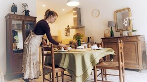 A person setting a table in a room with a wooden cabinet, a clock, and a mirror on the wall. - Maison des Genets (N°6) (Marcorignan)