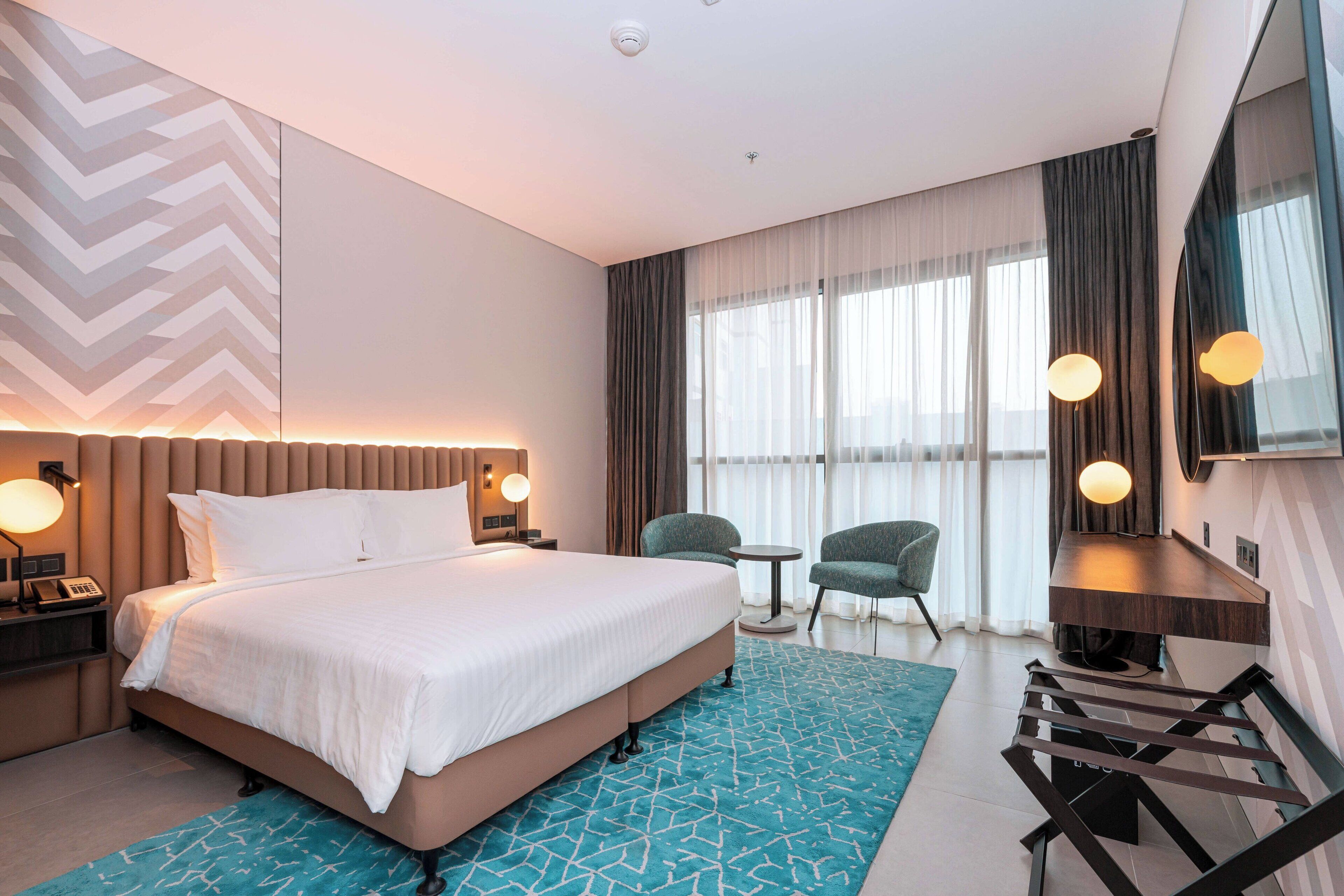 A modern hotel room with a large bed, a desk, two chairs, a small table, and a large window with curtains.