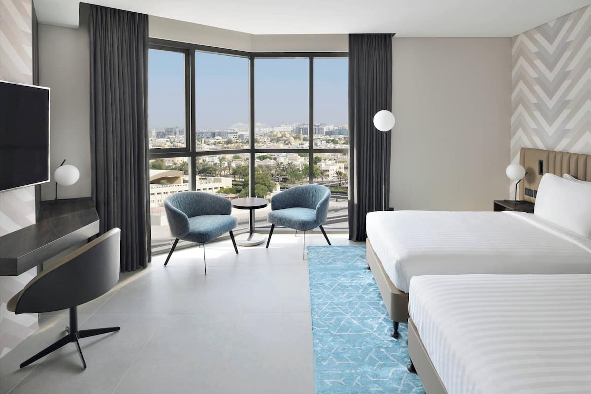Une chambre d’hôtel avec une grande fenêtre, deux fauteuils, un lit, un bureau et une télévision.