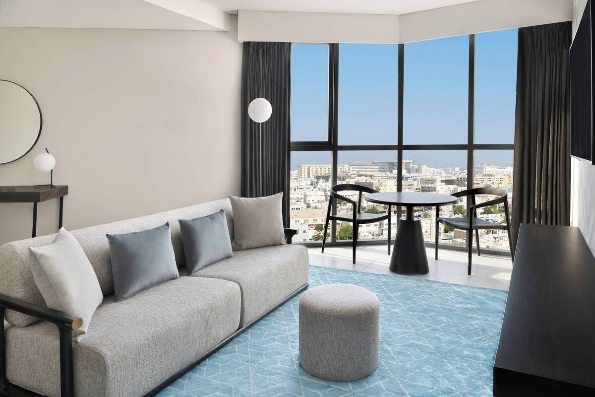 Un salon moderne avec un canapé gris, un pouf rond, une petite table et une vue sur la ville grâce à de grandes fenêtres.
