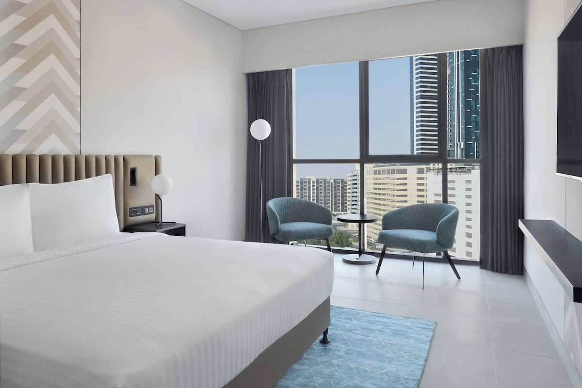 Une chambre d’hôtel moderne avec un grand lit, deux fauteuils, une petite table et une vue sur de hauts immeubles, visible à travers de grandes fenêtres.