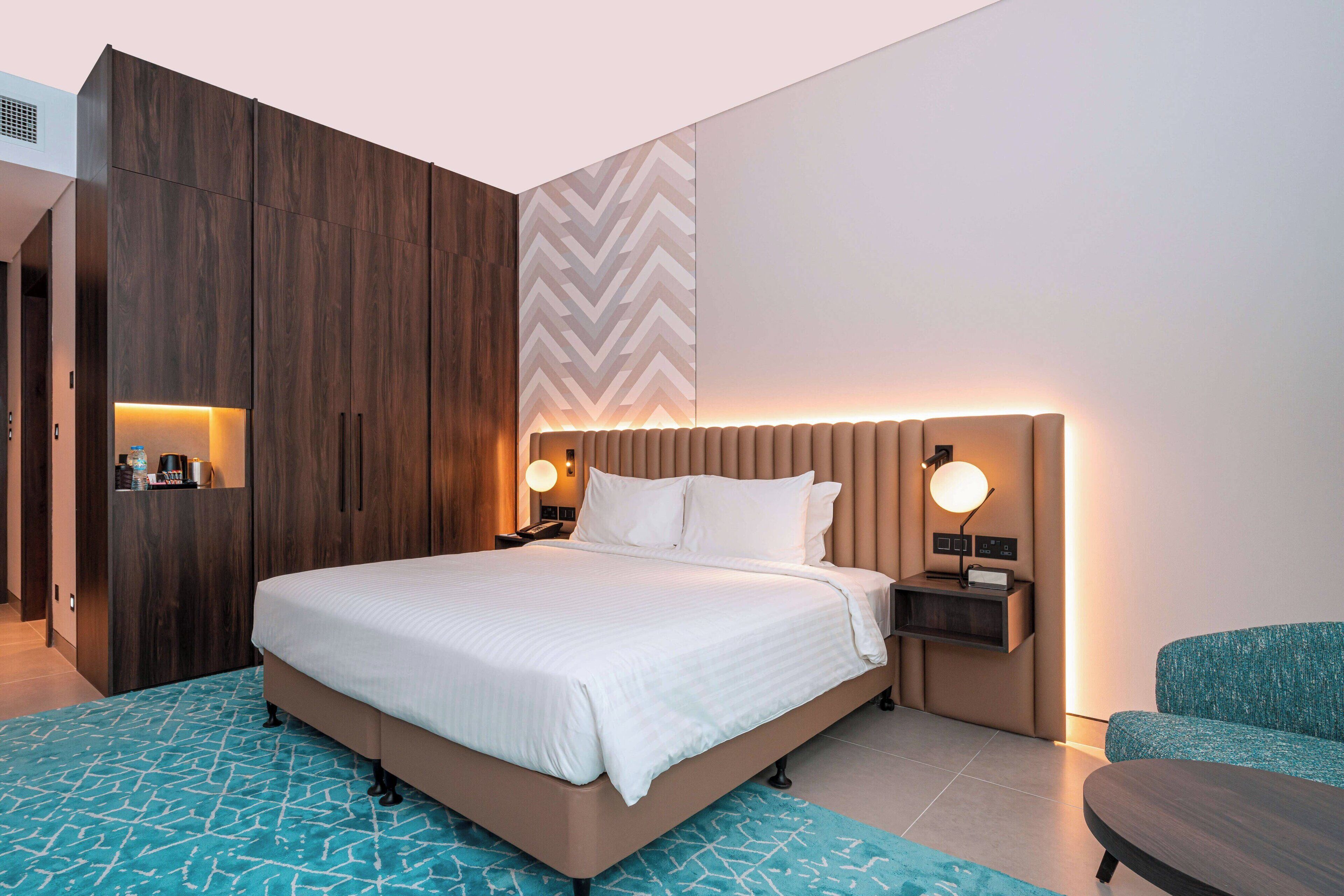 A modern hotel room with a large bed, a nightstand, a seating area, and a wardrobe.