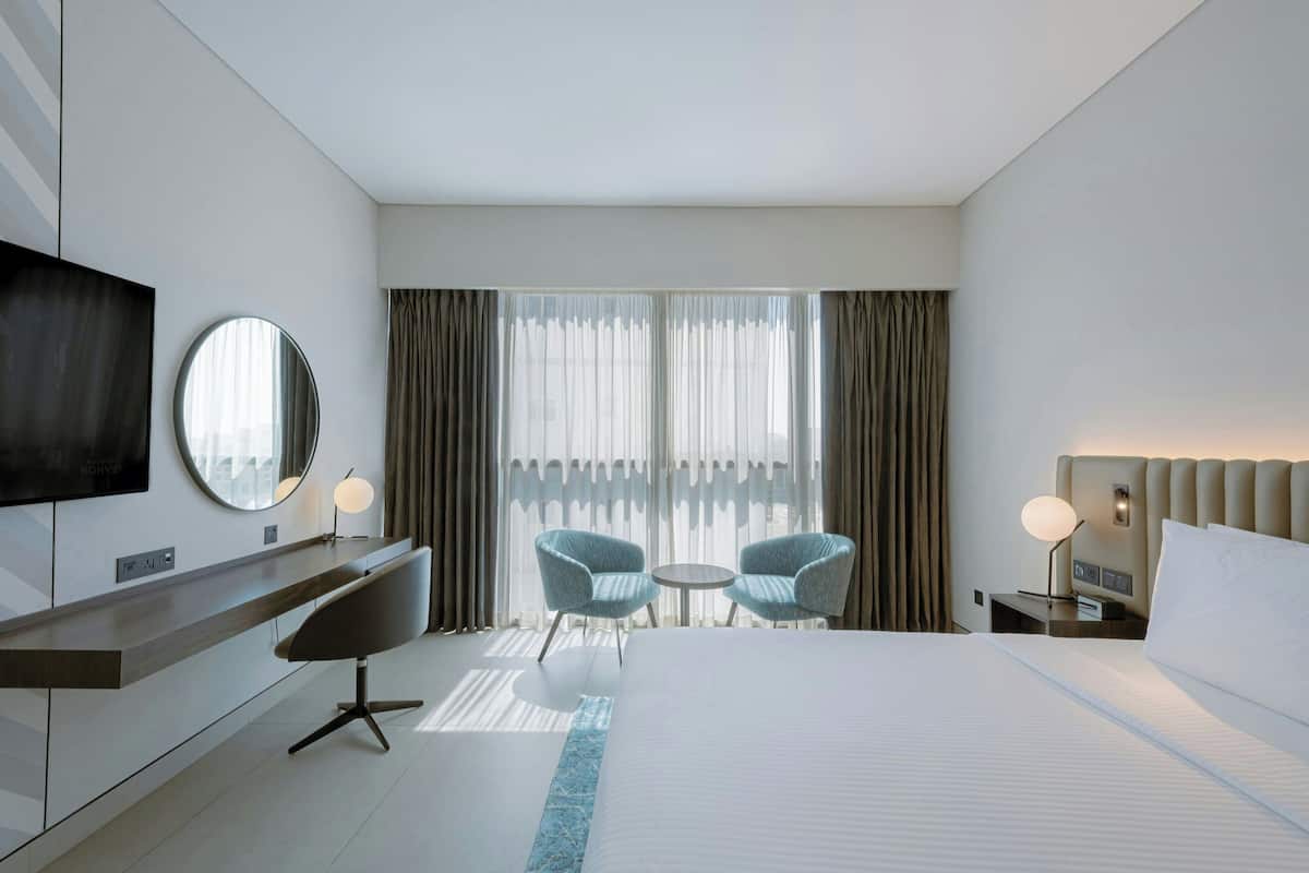 Une chambre d’hôtel moderne dotée d’un grand lit, d’un bureau avec un téléviseur à écran plat, de deux fauteuils, d’un miroir rond et offrant une vue sur la ville à travers des rideaux transparents.
