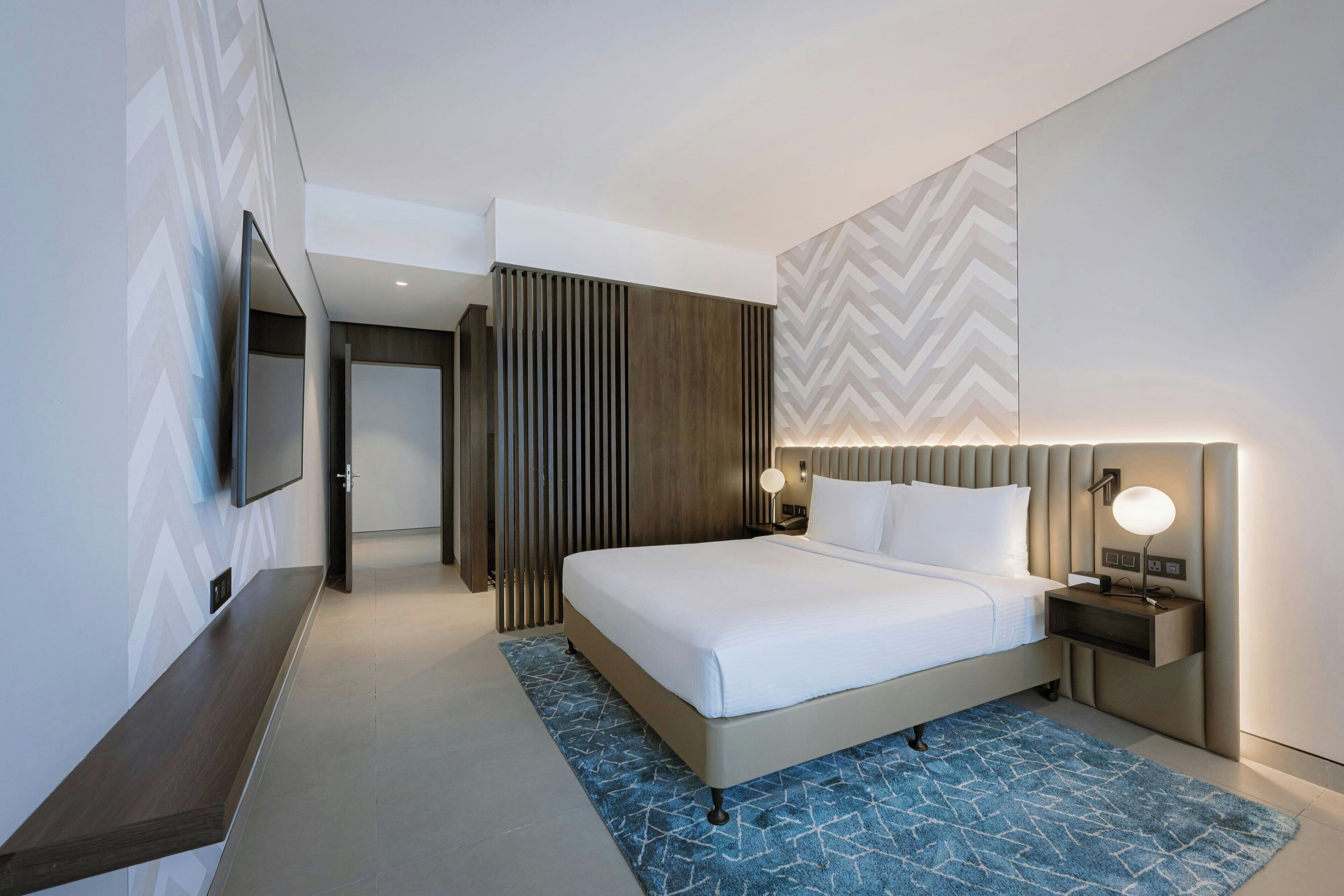 A modern hotel room with a large bed, a flat-screen TV mounted on the wall, a bedside table with a lamp, and a patterned rug.