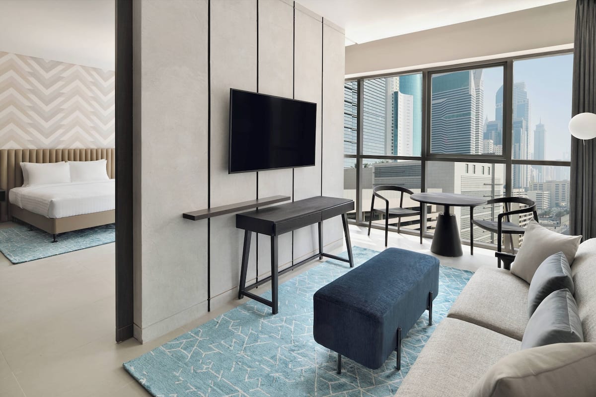 Une chambre d’hôtel moderne, équipée d’un lit, d’un téléviseur fixé au mur, d’un canapé, d’une petite table et offrant une vue sur le paysage urbain grâce à de grandes fenêtres.
